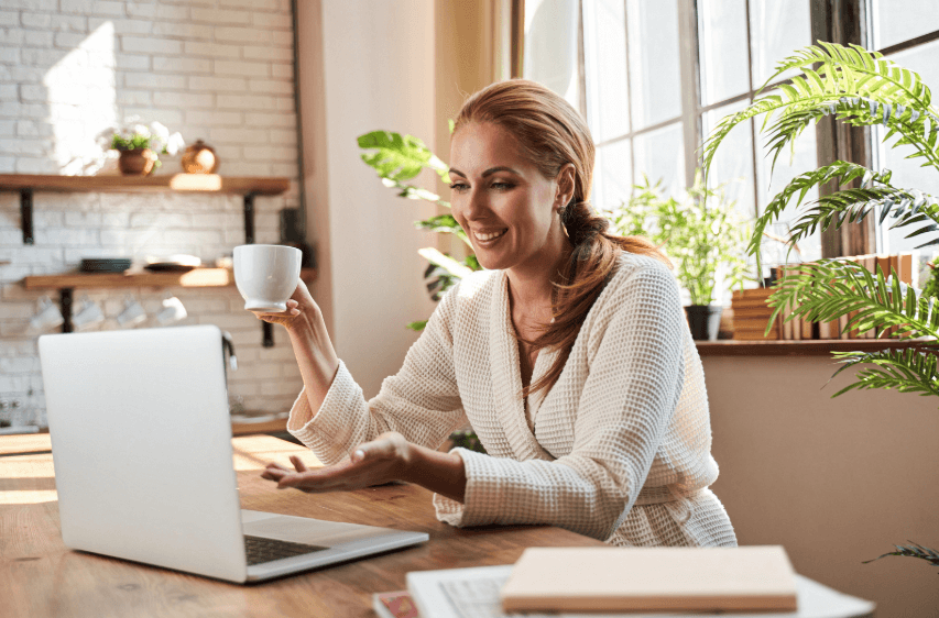 A photo of woman sitting with a white coffee cup in her hand, while looking at a computer screen and smiling.