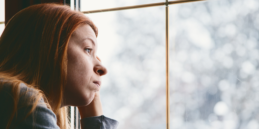 Woman with red hair staring out a window to a snowy yard, coping with seasonal depression.