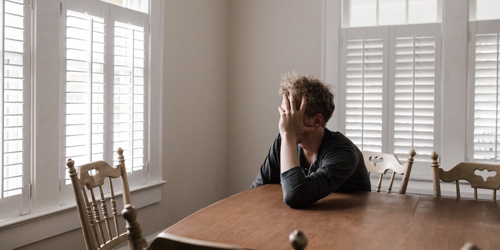 A man sitting anxiously at a kitchen table with his head in his hand. He is thinking negative thoughts.