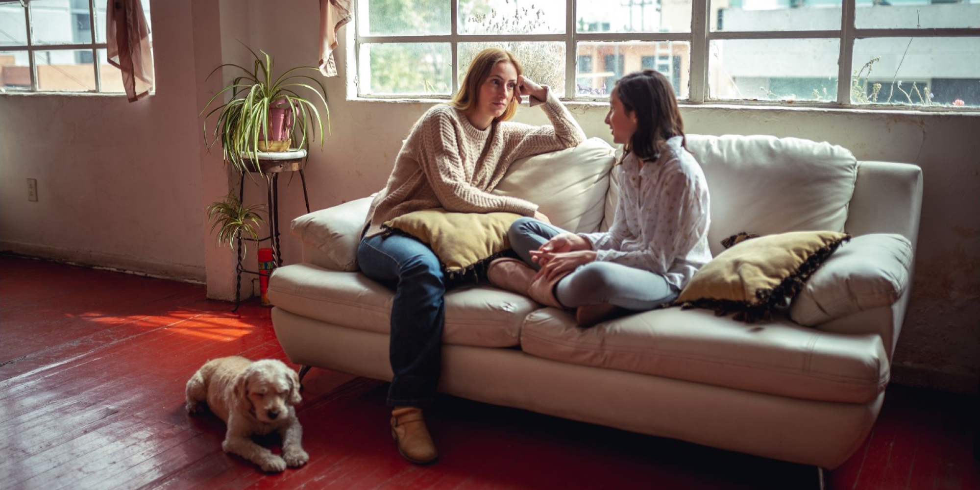 A mother and daughter sit. on a couch in front of windows. A dog sits on the floor in front of them. They look to be having a serious conversation.