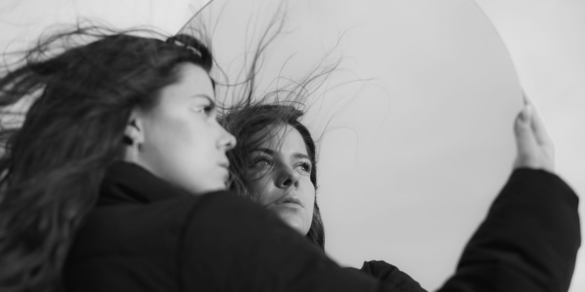 A black and white photo of a woman holding a mirror close to her face, while her hair is blowing in the wind.