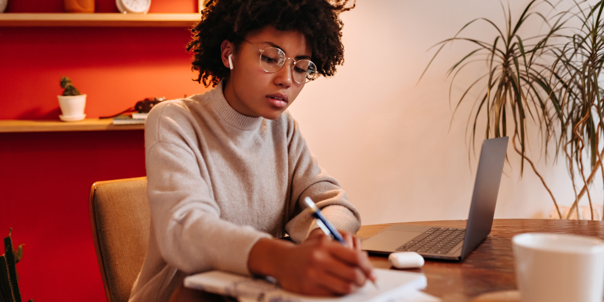 A woman sitting against an orange accent wall, listening to her AirPods and writing in her journal.