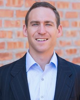 A man in a dress shirt and suit jacket standing against a brick wall.