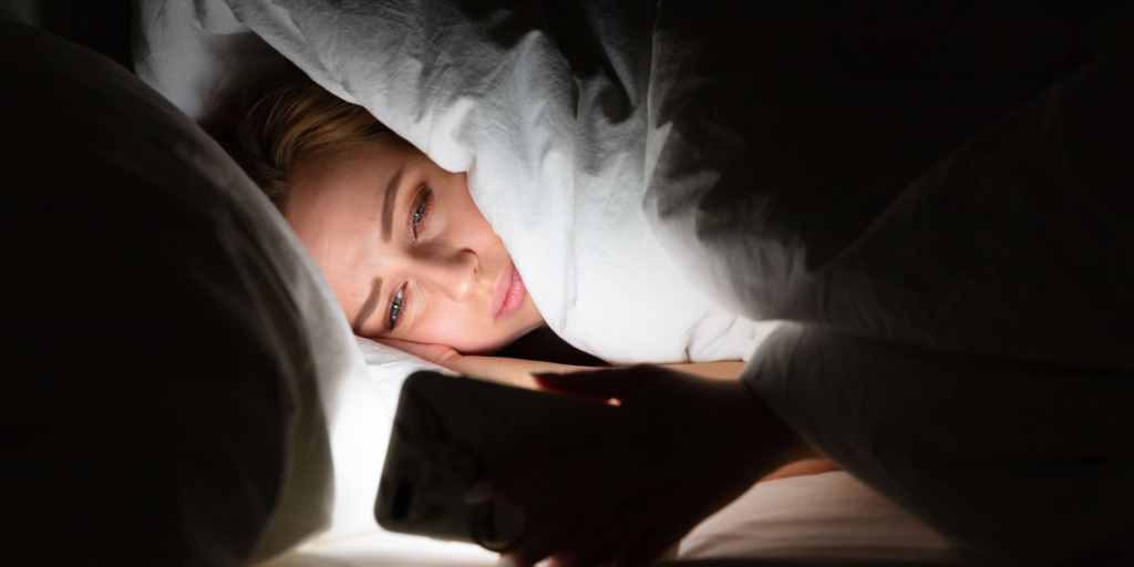 A woman lying under the covers and scrolling on her phone. She is experiencing revenge bedtime procrastination